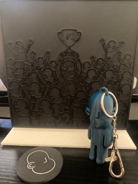 The image features a small blue keychain attached to a larger black display board with a picture of a crowd of people on it. The keychain has a silver chain hanging from it. In front of the display, there's also a coaster featuring an image of a person's head and a logo for @npconchain.