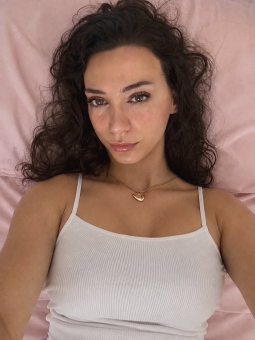 A selfie-style portrait of a young woman with long curly dark hair, wearing a white ribbed tank top and a delicate gold heart necklace, lying against pink satin bedding.