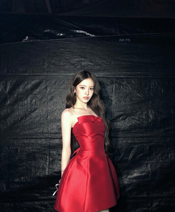 A photo of a woman with long dark wavy hair wearing a red strapless dress with a bow detail, posed against a black backdrop.