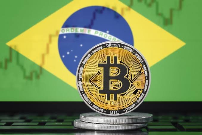 A physical Bitcoin token with the words "bitcoin," "BTC," "secure," and "digital" inscribed on its edge sits atop a stack of coins, with the Brazilian flag blurred in the background. The image illustrates Brazil's new crypto transfer regulation set to take effect on January 1, 2027, which will impose up to a 24-hour review period on transfers exceeding $10,000 from Brazilian exchanges to foreign platforms or self-custody wallets in an effort to prevent fraud.