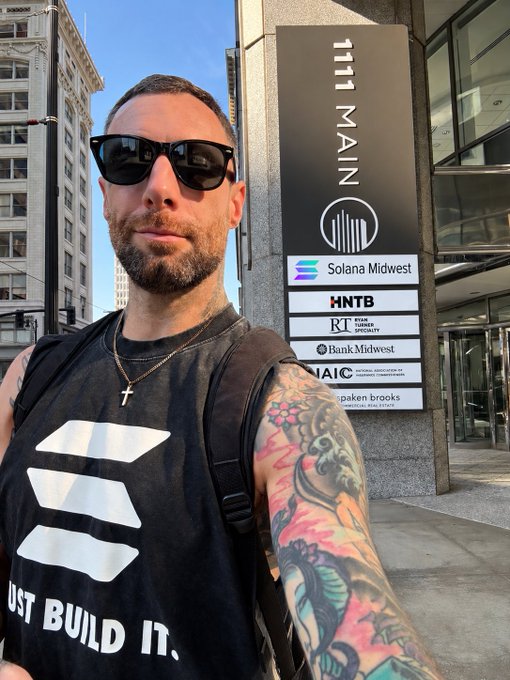 A bearded man wearing sunglasses and a black 'JUST BUILD IT.' tank top poses for a selfie in front of a building directory sign reading '1111 MAIN' with tenants including Solana Midwest, HNTB, and Bank Midwest.