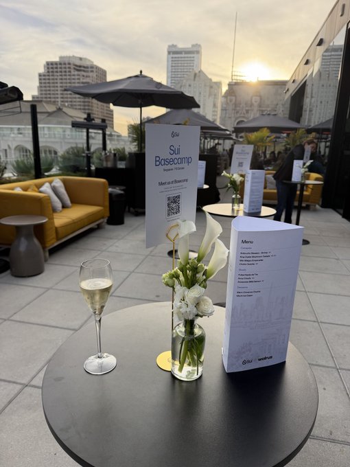 A rooftop event setup with a table holding a champagne glass, a floral arrangement of calla lilies, and signs reading 'Sui Basecamp' and 'Menu', with the Sydney Opera House visible in the background.