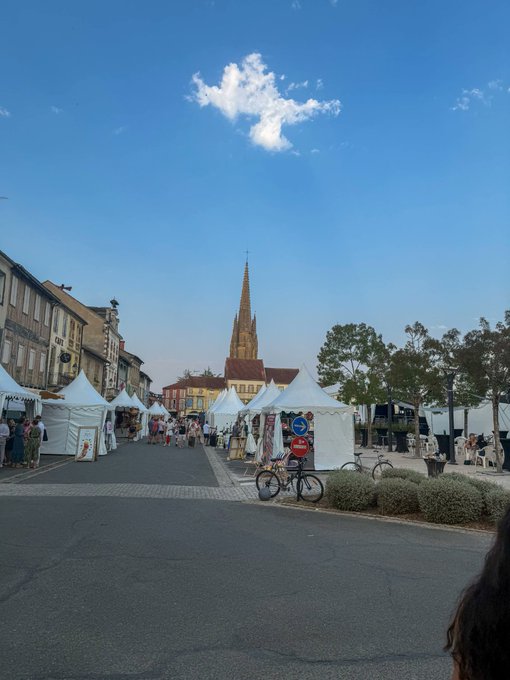 An outdoor market scene with white tents lining a street, with a gothic church spire rising in the background under a blue sky.