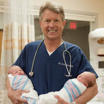 A smiling doctor wearing blue scrubs and a stethoscope, holding two newborn babies wrapped in striped hospital blankets in what appears to be a hospital room.