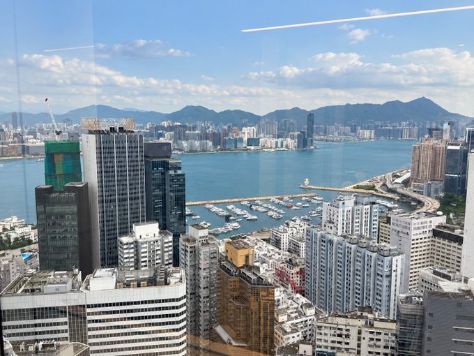 An elevated cityscape view, likely from a high-rise window, overlooking a dense urban area with a harbor, marina, bridges, and mountains in the distance under a partly cloudy sky.