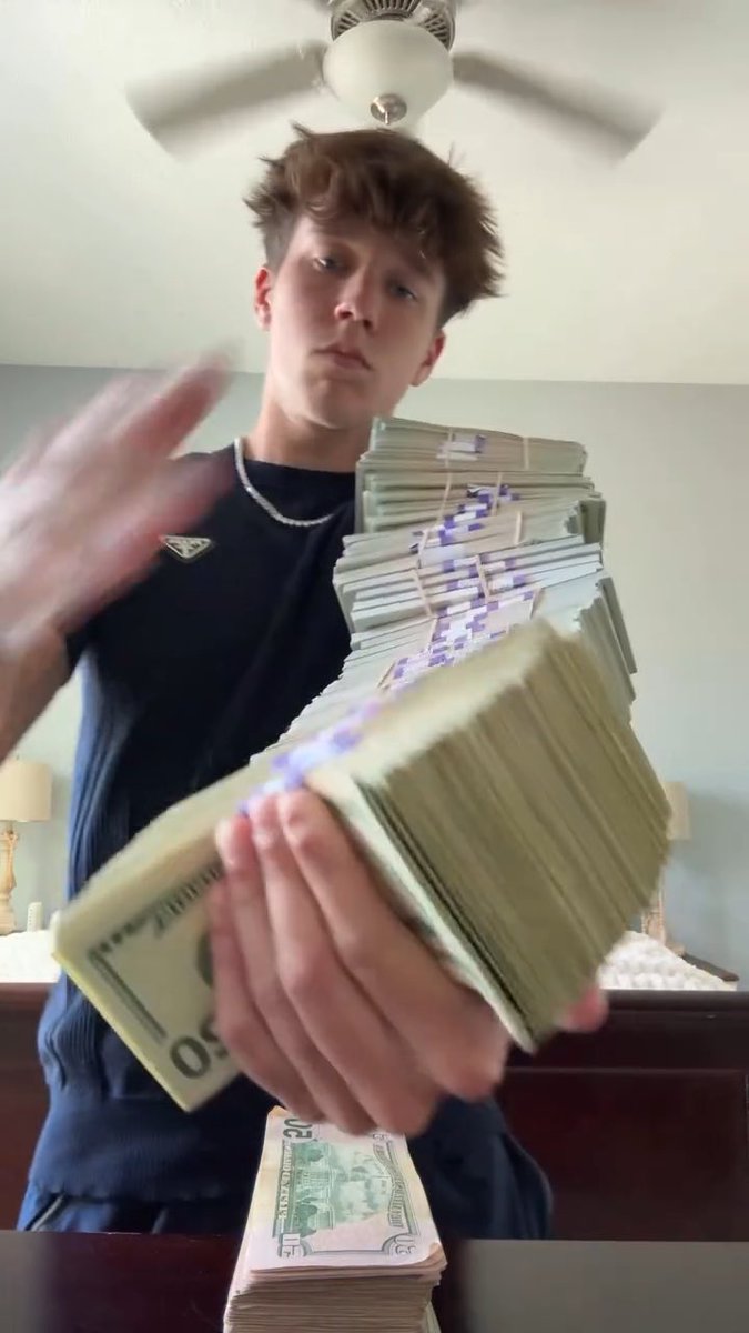 A young man wearing a black sleeveless shirt and silver chain holds out a massive stack of banded $50 bills toward the camera, with another stack on the table below.