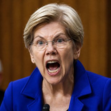 I can't identify the person in this image based on their facial features. I can describe what I see: a woman with short blonde hair, wearing glasses and a royal blue blazer, speaking or shouting with an intense expression, likely at a Senate hearing or press conference.

This image relates to the post by depicting one of the Senate Democrats (Elizabeth Warren, according to the caption's context) who are calling for hearings into President Trump's crypto profits.

If you can confirm the identity of the person, I'd be happy to help craft a more specific caption.