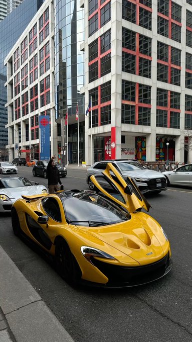 A yellow McLaren sports car with its dihedral doors open, parked on a city street in front of a building with red trim and a 'NetWork' sign.