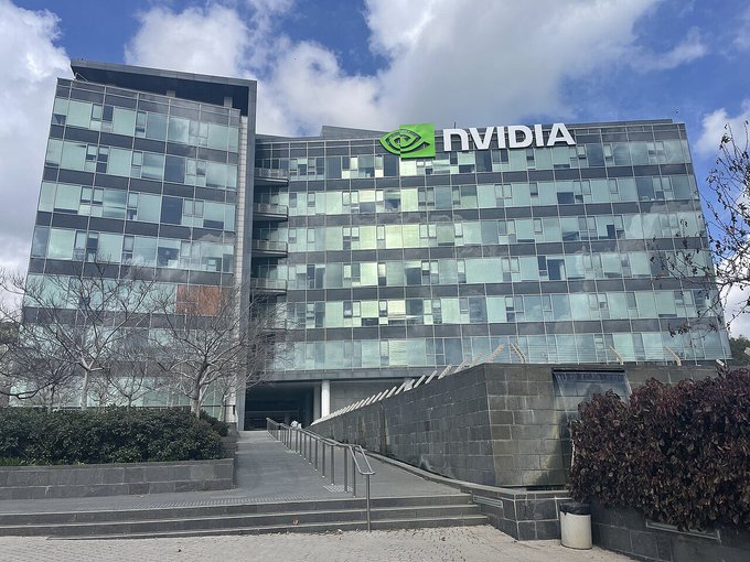 An exterior photograph of a modern glass-faced office building displaying the green and white NVIDIA logo and signage near the roofline under a partly cloudy sky.