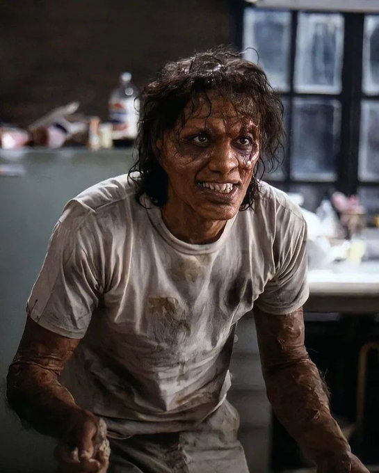 A still image of a distressed woman with darkened, disheveled hair, dirt-covered skin, and bared teeth, wearing a stained white t-shirt in a dimly lit, messy interior.