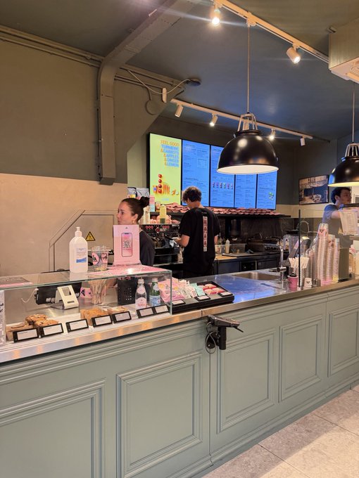 A cozy café interior with a pastel-green service counter displaying pastries under glass, illuminated by black pendant lights, while baristas prepare orders near a digital menu board. The "Good morning" greeting pairs perfectly with the warm, welcoming scene of a fresh Sunday coffee stop.
