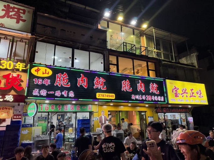 A bustling nighttime street scene in China featuring brightly lit storefronts with red Chinese characters, including signs for '脆肉鲩' and '宝兴正宗', with a crowd of people dining below.