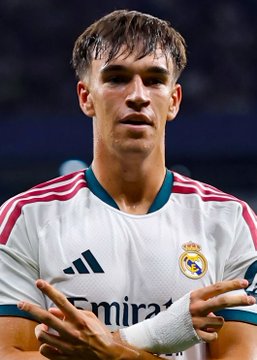 A young soccer player wearing a white Real Madrid jersey with pink and teal accents celebrates with his hands crossed.