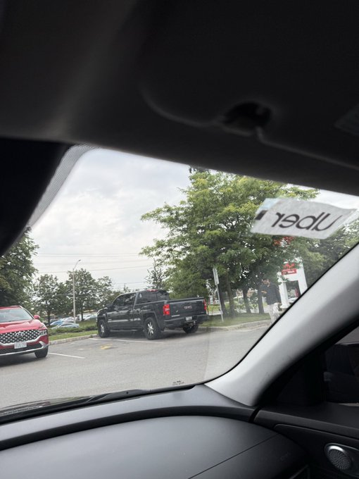 A view from inside a car looking out the windshield at a parking lot, showing a black Ford pickup truck parked near a red SUV, with trees and a commercial area visible in the background.