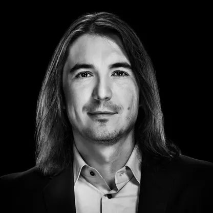 A black-and-white portrait shows a man with long dark hair, a light mustache and goatee, wearing a dark suit jacket over a white collared shirt, smiling slightly against a black background. The image appears to be a professional headshot of Vladimir Tenev, co-founder and CEO of Robinhood, relating to the post about the trading platform's record-breaking $8 million in single-day revenue.