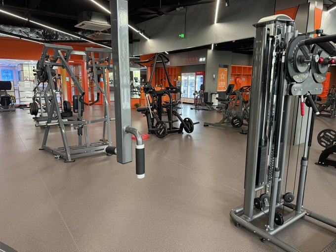 An indoor gym with weight machines, cable equipment, and orange accents on the walls, featuring a polished floor and bright overhead lighting.