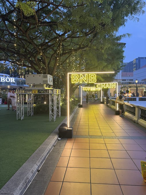 A glowing neon archway reading "BNB CHAIN BOOTH CHAMPIONSHIP" marks the entrance to an immersive crypto event venue, where string lights drape from the trees above and interactive project booths line the tiled walkway. The lively atmosphere and scattered attendees capture the energetic vibe described in the post, where visitors explore ecosystem booths and collect badges for a chance to win prizes like BNB sneakers.