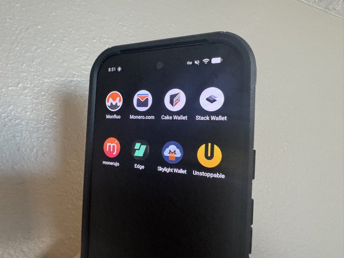 A smartphone with a rugged black case displays the home screen with a folder containing various cryptocurrency wallet apps, including Monfluo, Monero.com, Cake Wallet, Stack Wallet, monerujo, Edge, Skyflight Wallet, and Unstoppable—likely apps being evaluated for an upcoming GrapheneOS Monero wallet review video.