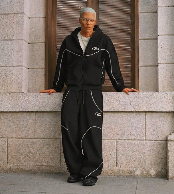 A person with a shaved head and glasses models a black tracksuit with white piping and a logo, standing in front of a wooden door on a stone wall.