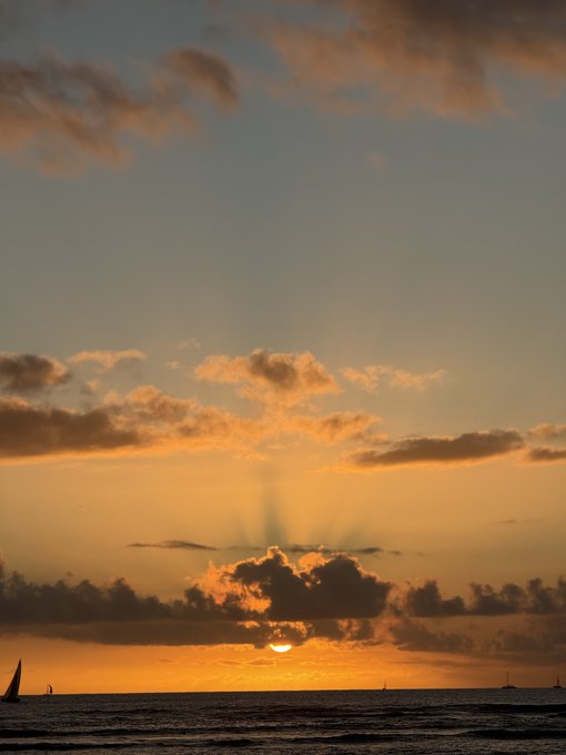 A serene sunset over the ocean with the sun partially hidden behind a cloud, casting golden rays through scattered clouds and silhouetting a small sailboat on the horizon.
