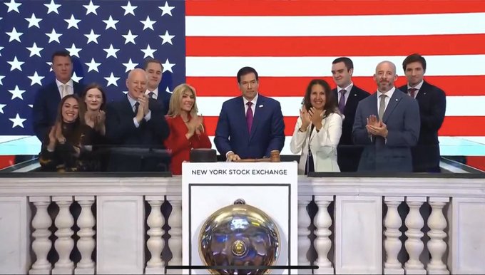 Secretary of State Marco Rubio stands at the New York Stock Exchange podium marked "NEW YORK STOCK EXCHANGE," preparing to ring the opening bell, surrounded by applauding officials and guests in front of a large American flag backdrop.