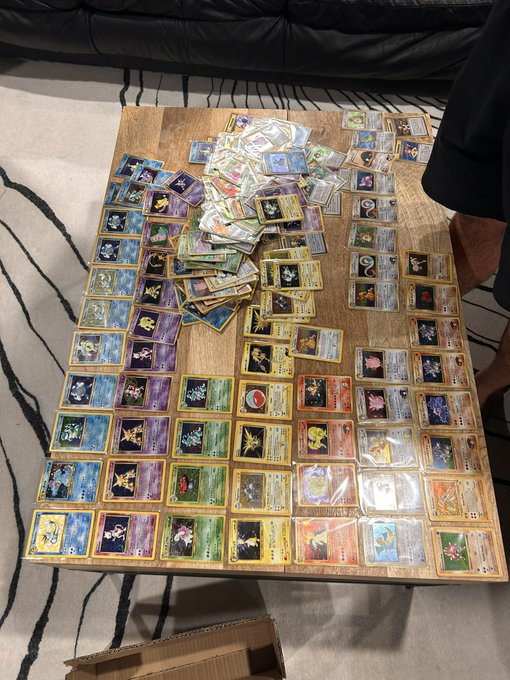 A large collection of Pokémon trading cards spread across a wooden table, neatly organized in rows with holographic and rare cards visible.