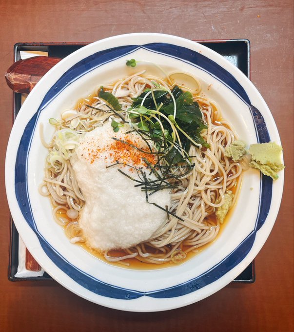A bowl of Japanese soba noodles topped with white fish, green herbs, seaweed strips, and a sprinkle of chili powder, served with wasabi on the side.