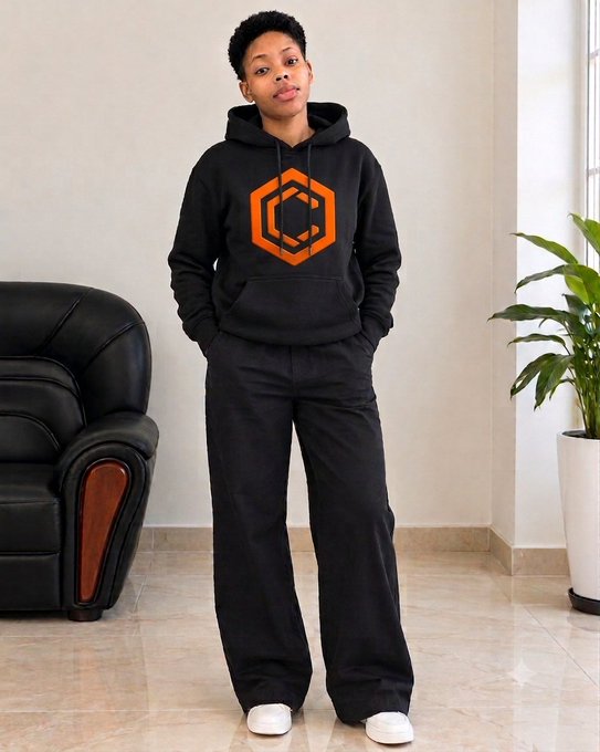 A young woman stands indoors wearing a black hoodie featuring an orange geometric hexagonal logo, paired with black pants and white sneakers.