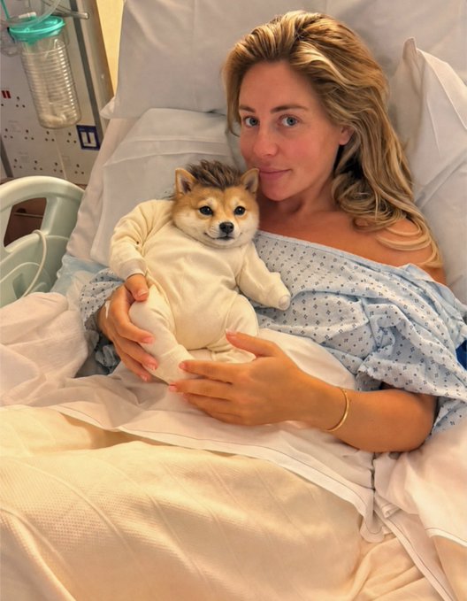A woman wearing a hospital gown lies in a hospital bed, holding and smiling alongside a small Shiba Inu dog dressed in a white outfit.