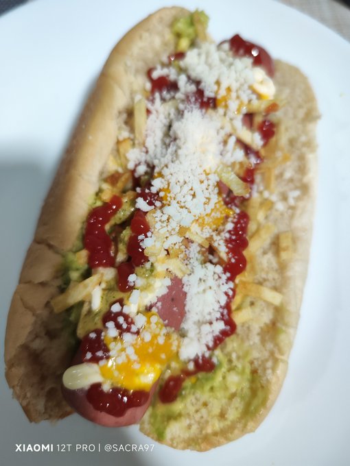 A loaded hot dog topped with ketchup, mustard, mayonnaise, guacamole, crispy potato sticks, and crumbled white cheese sits on a white plate, perfectly matching the "Perro caliente para cenar #hive" caption of a hot dog dinner. The image bears a "XIAOMI 12T PRO | @SACRA97" watermark in the corner.