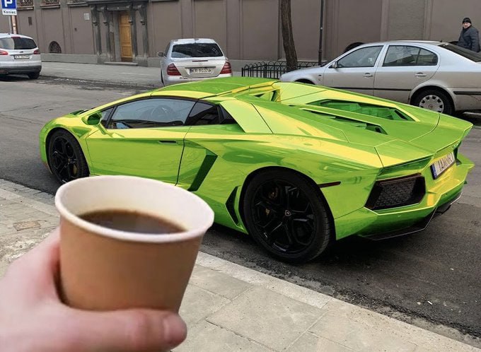 A hand holding a paper cup of coffee in the foreground, with a bright green/chrome Lamborghini parked on the street in the background.