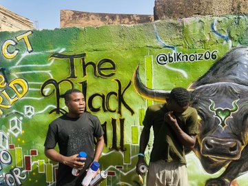Two young men stand in front of a green graffiti wall featuring a large bull design with text 'The Black Bull' and the handle '@bknoiz06'.