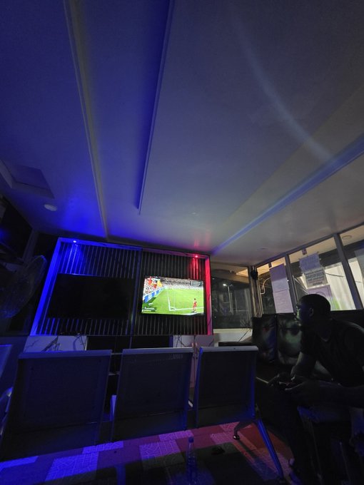 A dimly lit room with blue and red ambient lighting where a person sits in a chair watching a soccer match on a wall-mounted television.