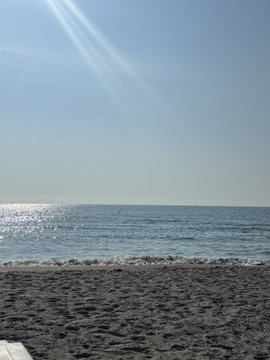 A serene view of a calm ocean from a sandy beach under a clear blue sky with subtle wispy clouds.