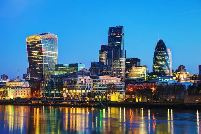 A nighttime cityscape of London featuring illuminated skyscrapers, including the Walkie-Talkie and Gherkin buildings, with their lights reflecting on the River Thames.