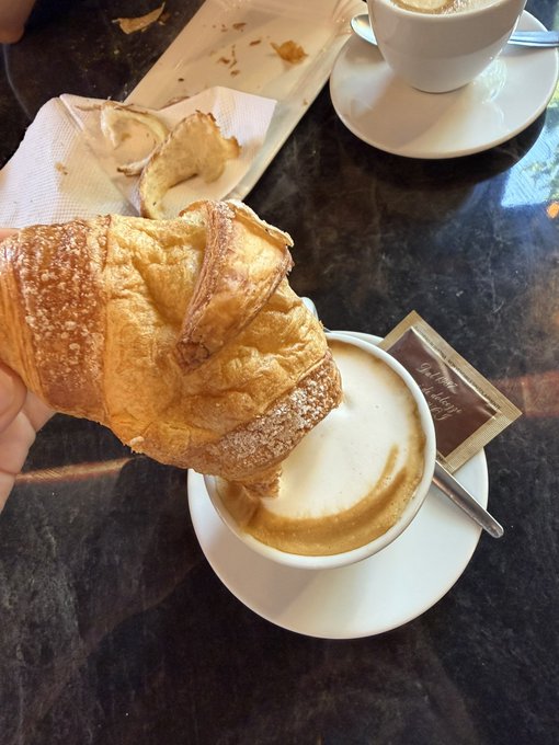 A hand holds a powdered sugar-dusted croissant above a cappuccino on a dark marble cafe table, with a coffee cup and saucer visible in the background.