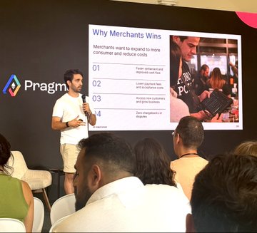 A man speaking at a podium during a presentation for 'Pragma', displaying a slide titled 'Why Merchants Wins' that explains merchants expand to more consumer and reduce costs through cash flow, product fees, and zero chargebacks.