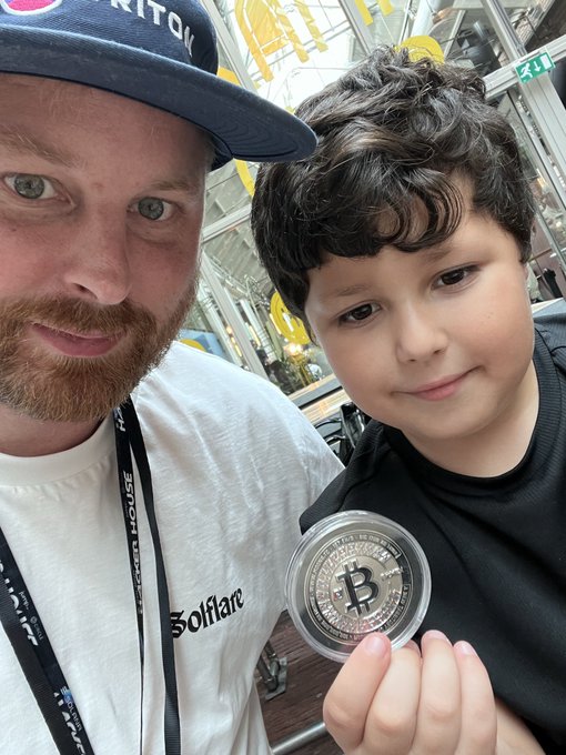 A selfie of a bearded man wearing a baseball cap and a white 'Bullfare' t-shirt alongside a young boy, with the boy proudly displaying a physical Bitcoin commemorative coin in his hand.