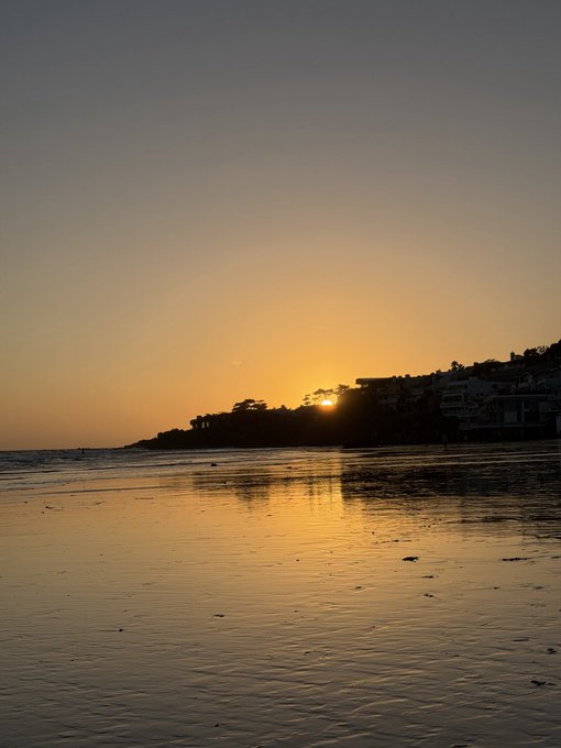 A serene sunset photograph captures the golden sun sinking behind a silhouetted coastal landscape, with its warm light reflecting beautifully across the wet sand of the shoreline.