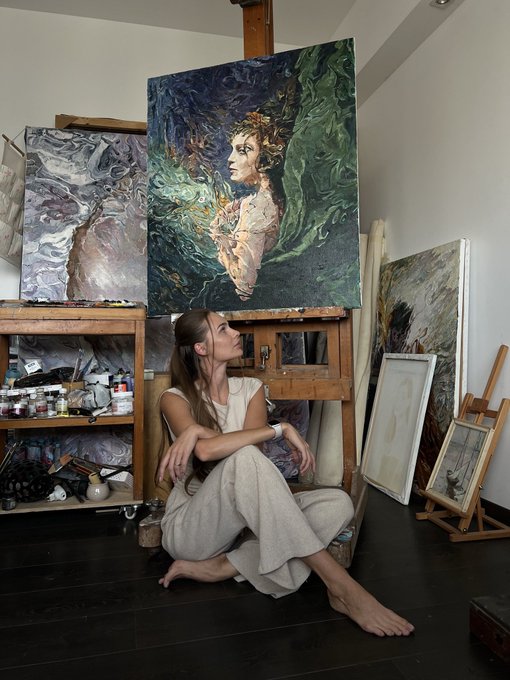 A woman sits barefoot on the floor of an art studio, surrounded by large, expressive oil paintings and art supplies.