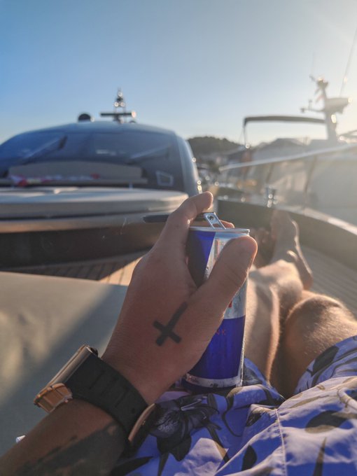 A first-person shot captures a hand adorned with a small "X" tattoo on the thumb, holding a blue and silver Red Bull can while lounging on a yacht with a yacht visible in the background. The luxury, leisurely setting complements the relaxed "Good morning Web3 fam, Have a fun hump day" vibe of the post.