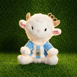The image features a stuffed animal, specifically a white goat plush toy with horns sitting on top of green grass. The background consists of a green carpet or lawn, which provides a contrasting backdrop for the white goat. There are no other objects or text present in the image.