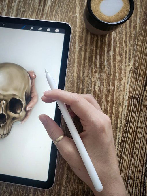 A hand holds a digital stylus over an iPad displaying a detailed illustration of a human skull, with a coffee mug resting nearby on a wooden surface — visually tying into the "Remains" NFT collection referenced in the post. The artist's workspace mirrors the macabre aesthetic of the artwork, complementing the announcement of a new collector unlocking a mint.
