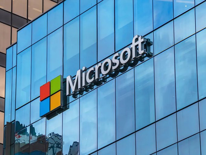 Microsoft corporate logo and name mounted on the glass facade of a modern office building, reflecting the sky.
