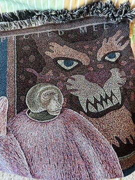 A close-up of a woven tapestry featuring a stylized cat face with blue eyes next to a small astronaut figure, with the partial text 'CIEO MEE' visible at the top.
