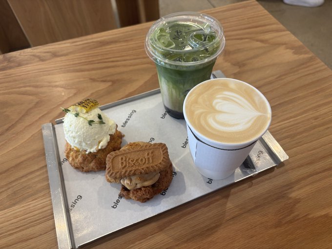 A café scene featuring a metal tray with a matcha latte, a regular latte with heart-shaped foam art, and two pastries including one labeled 'Bisou,' set on a wooden table.
