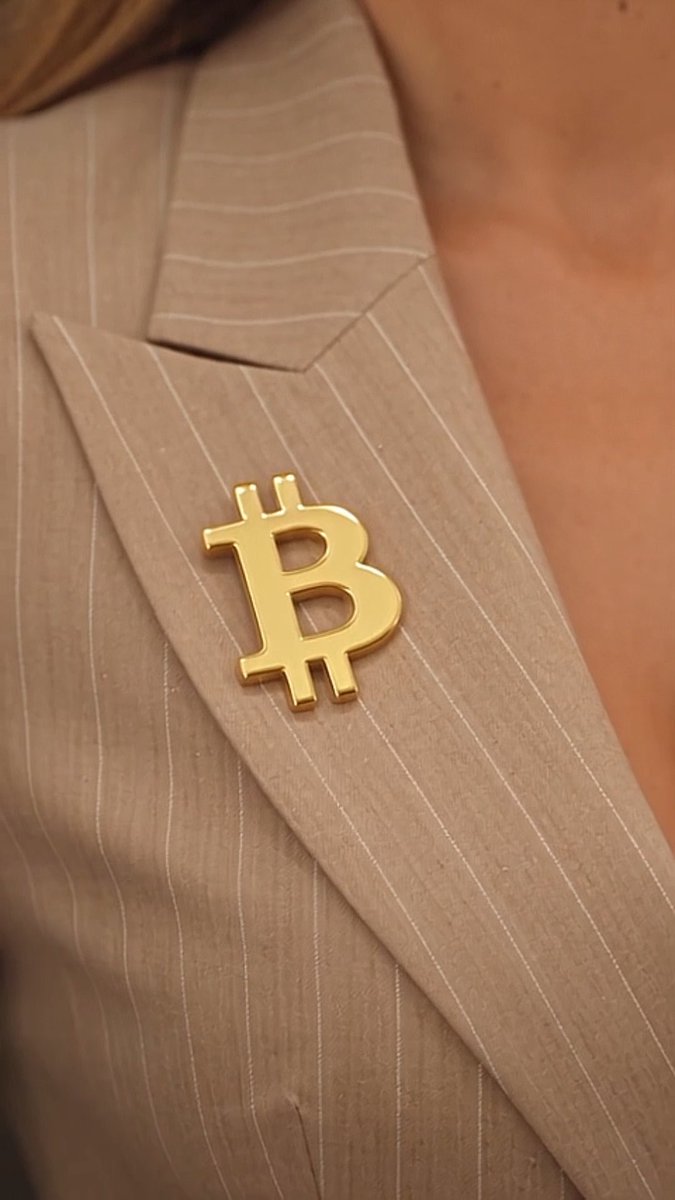 Close-up of a gold Bitcoin (₿) lapel pin worn on a beige pinstripe suit jacket.