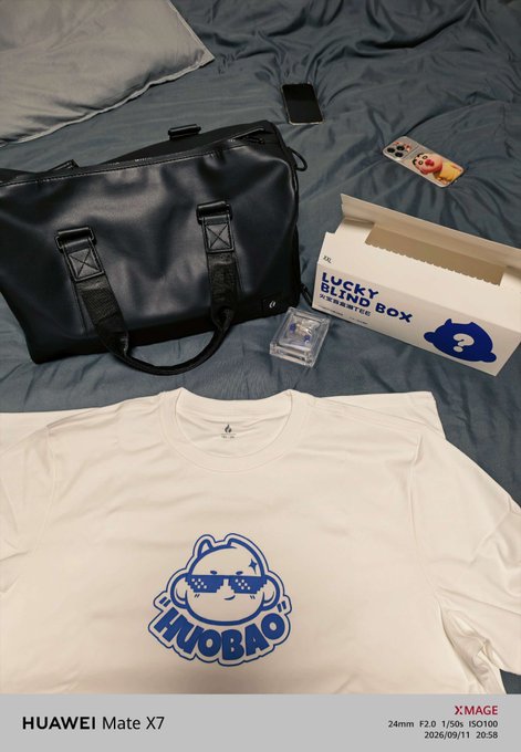 A HUAWEI Mate X7 phone displays an unboxing photo showing HTX (Huobi) 13th anniversary merchandise, including a black duffel bag, a "LUCKY BLIND BOX" featuring a blue cat mascot with pixel sunglasses labeled "HUOBAO," and a matching white long-sleeve shirt with the same cat design. The caption celebrates receiving the anniversary merch, with the "HuoBao" character representing HTX's journey through bull and bear markets over 13 years in the crypto industry.