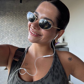 A smiling woman takes a selfie wearing round reflective sunglasses and wired white earphones.