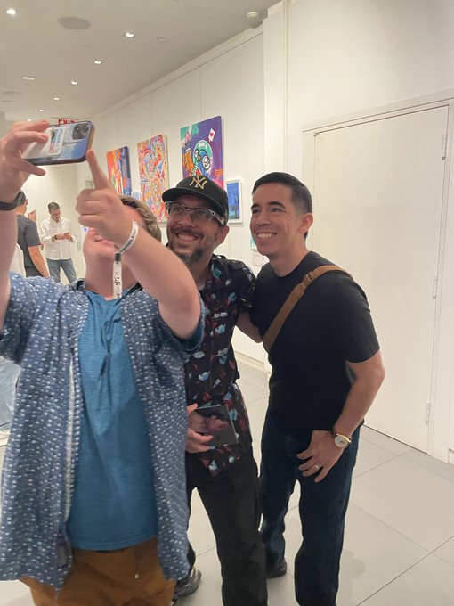 Three men smile while taking a selfie in what appears to be an art gallery, with colorful paintings visible on the white walls behind them.
